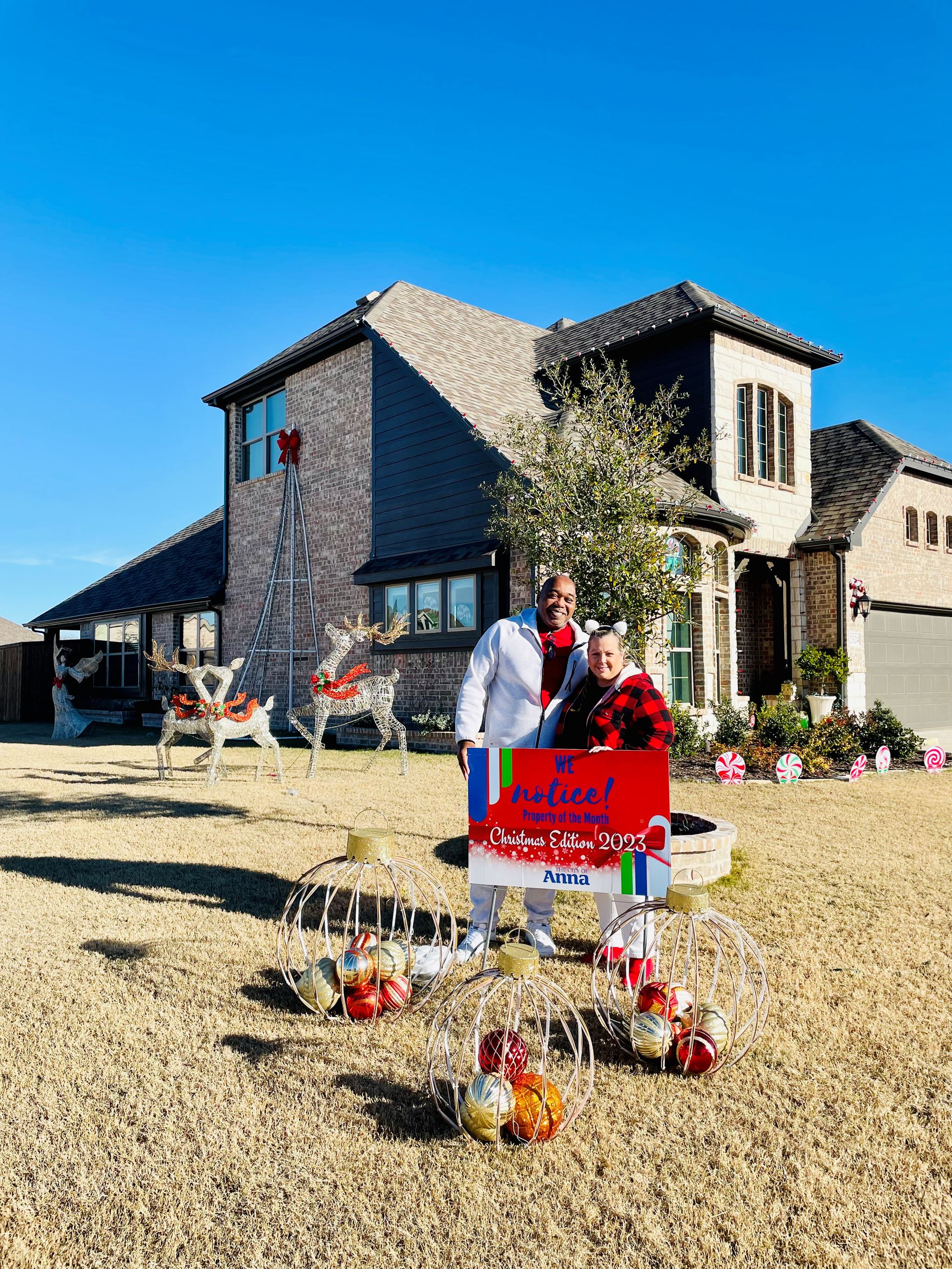 Man and woman standing in front of a house with Christmas edition "Property of the Month" yard
